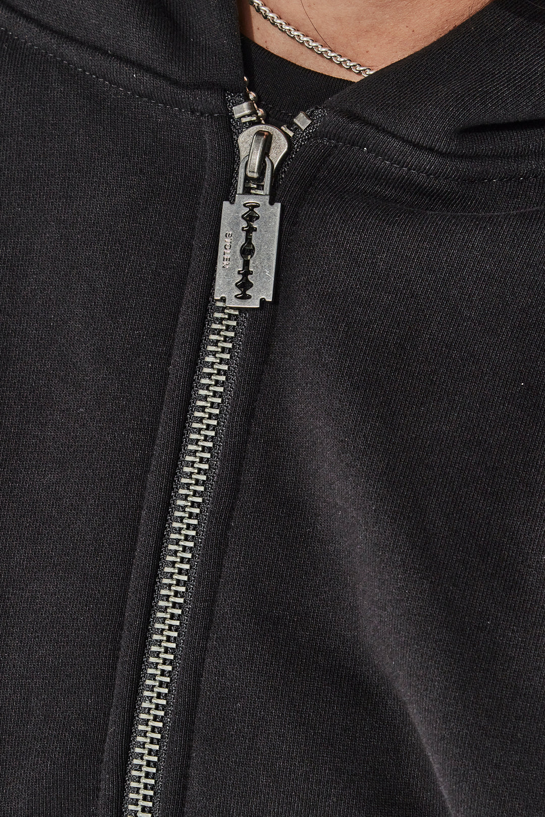 Close-up of a black zip-up hoodie with a razor blade design on the zipper pull.