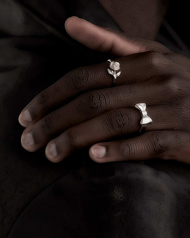 Stem Rose Ring styled on male model’s hand, shown close-up with Bow Ring, highlighting contrast in sculptural details against skin tone and natural light